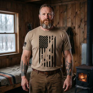 Bearded man with forest and elk tattoos standing in a rustic wood-paneled deer camp cabin by a wood stove, wearing the 2A American Flag T-Shirt in Brown Savana: distressed black American flag with an AR-15 rifle silhouette cut through the stripes on the front, American flag on the right sleeve