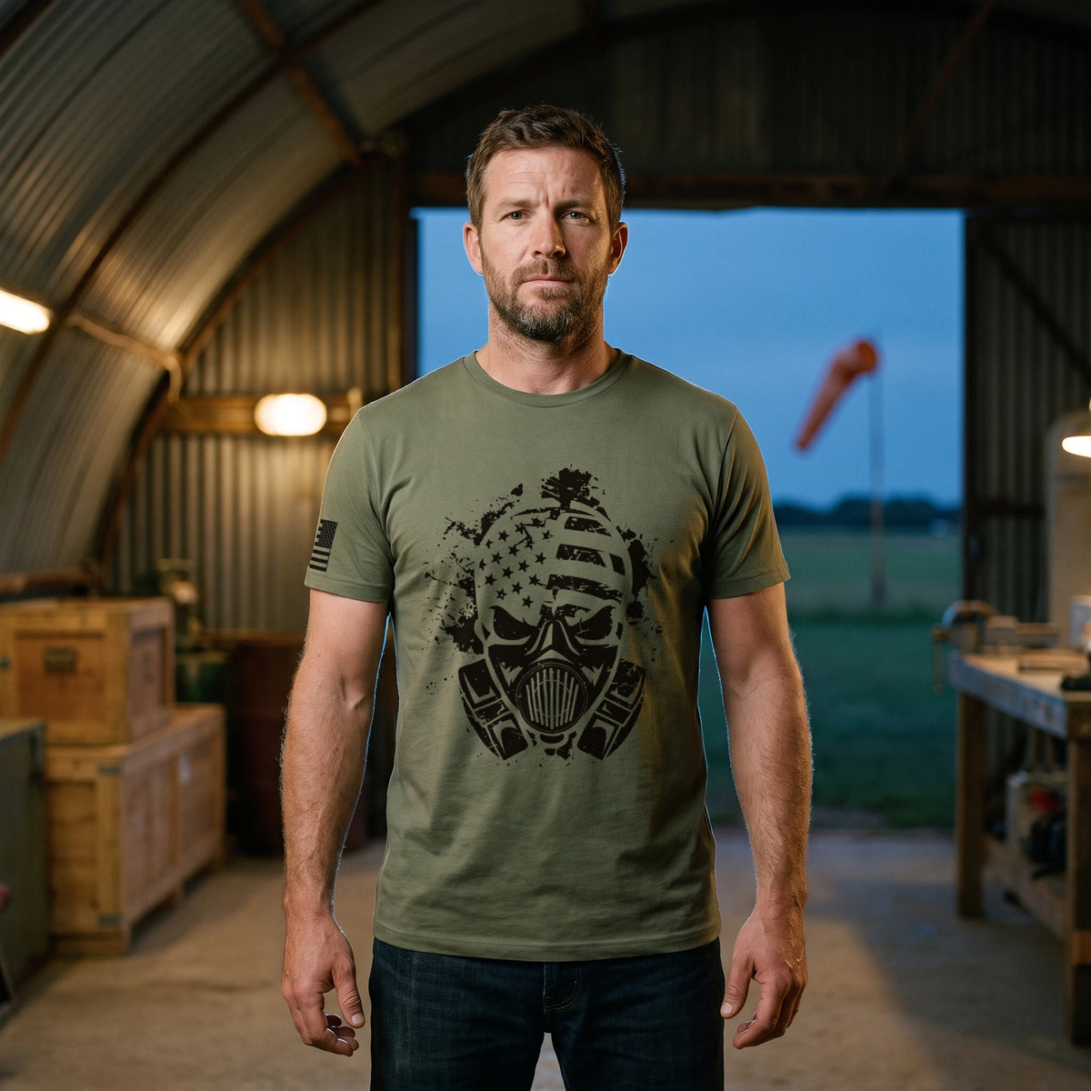Man standing in the open doorway of an old Quonset hut hangar on a grass airfield at dusk, wearing the American Fighter Skull T-Shirt in Military Green with the black gas mask skull printed large on the front and a flag on the right sleeve