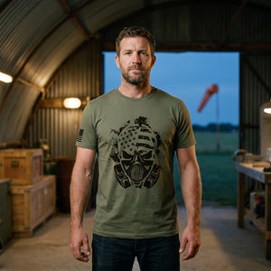 Man standing in the open doorway of an old Quonset hut hangar on a grass airfield at dusk, wearing the American Fighter Skull T-Shirt in Military Green with the black gas mask skull printed large on the front and a flag on the right sleeve
