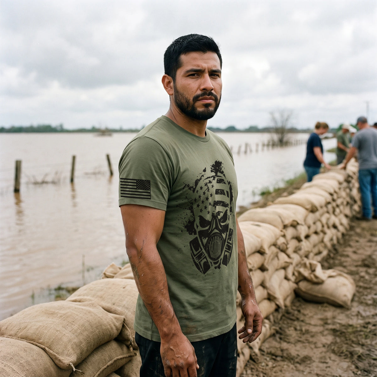 Man beside a wall of sandbags on a flooded river levee, turned to the side, wearing the American Fighter Skull T-Shirt in Military Green with the black gas mask skull printed large on the front and a flag on the right sleeve