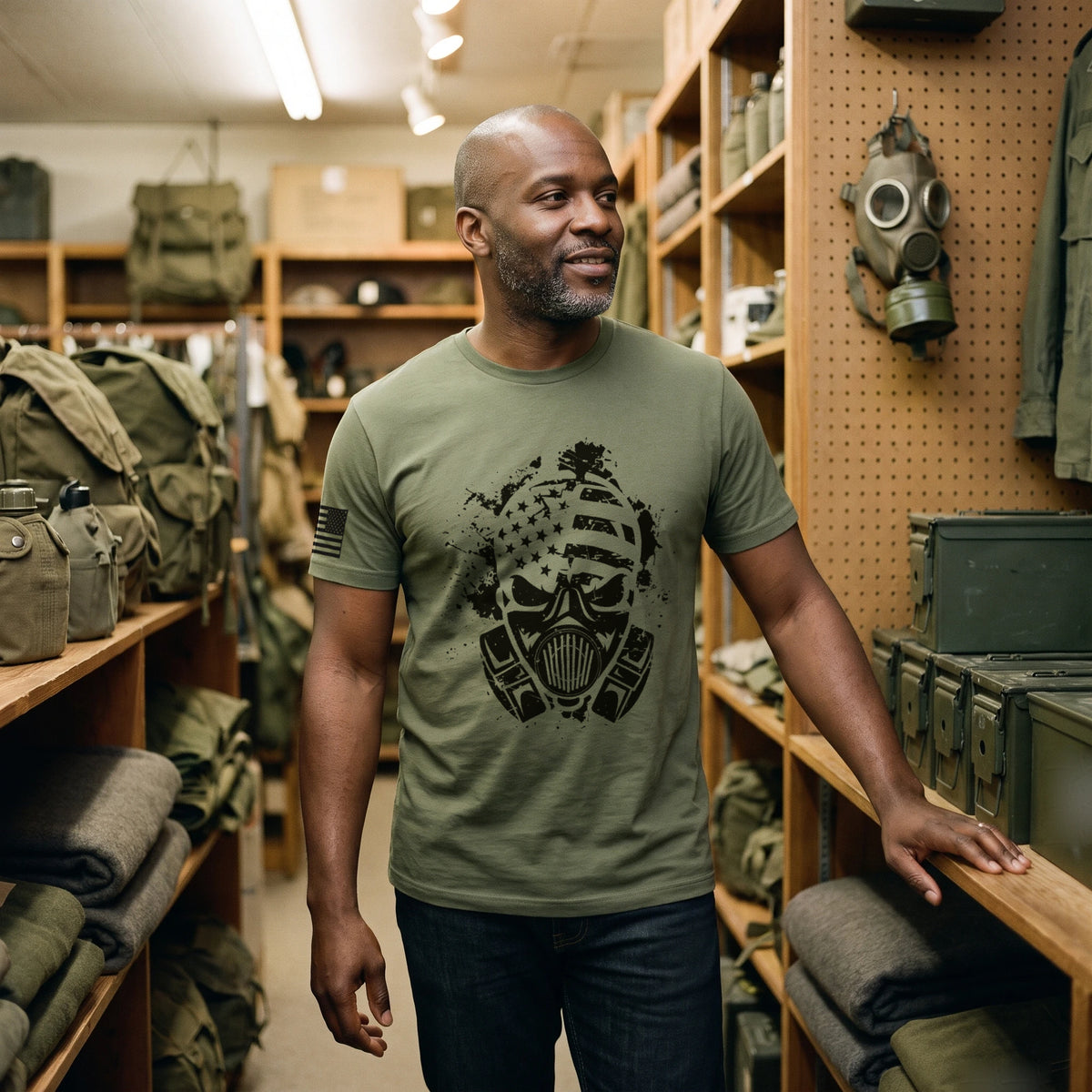 Man walking through an army-navy surplus store past canvas packs and a vintage gas mask hanging on the wall, wearing the American Fighter Skull T-Shirt in Military Green with the black gas mask skull printed large on the front