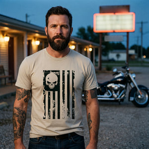 Man standing in the gravel lot of a roadside motor-court motel at blue hour with a black motorcycle behind him, wearing the American Flag Skull T-Shirt in Sand with the black skull flag printed large on the front