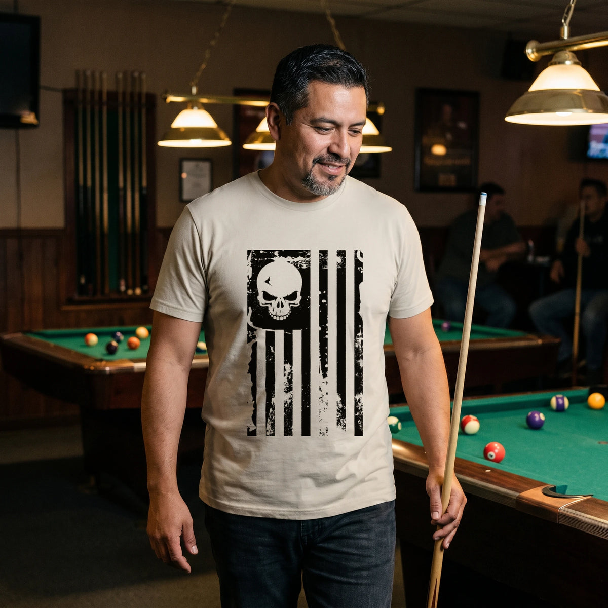 Man walking around a pool table in a pool hall with a cue at his side, wearing the American Flag Skull T-Shirt in Sand with the black skull flag printed large on the front