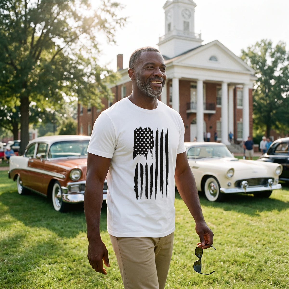 Man walking across a courthouse lawn between classic cars at a small-town car show, sunglasses in hand, wearing the American Flag T-Shirt in White with the tall vertical black brush-stroke American flag printed down the front