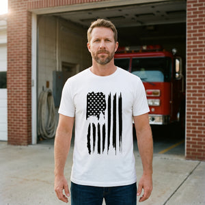 Man standing in front of the open bay doors of a small-town fire station with a red fire engine behind him, wearing the American Flag T-Shirt in White with the tall vertical black brush-stroke American flag printed down the front