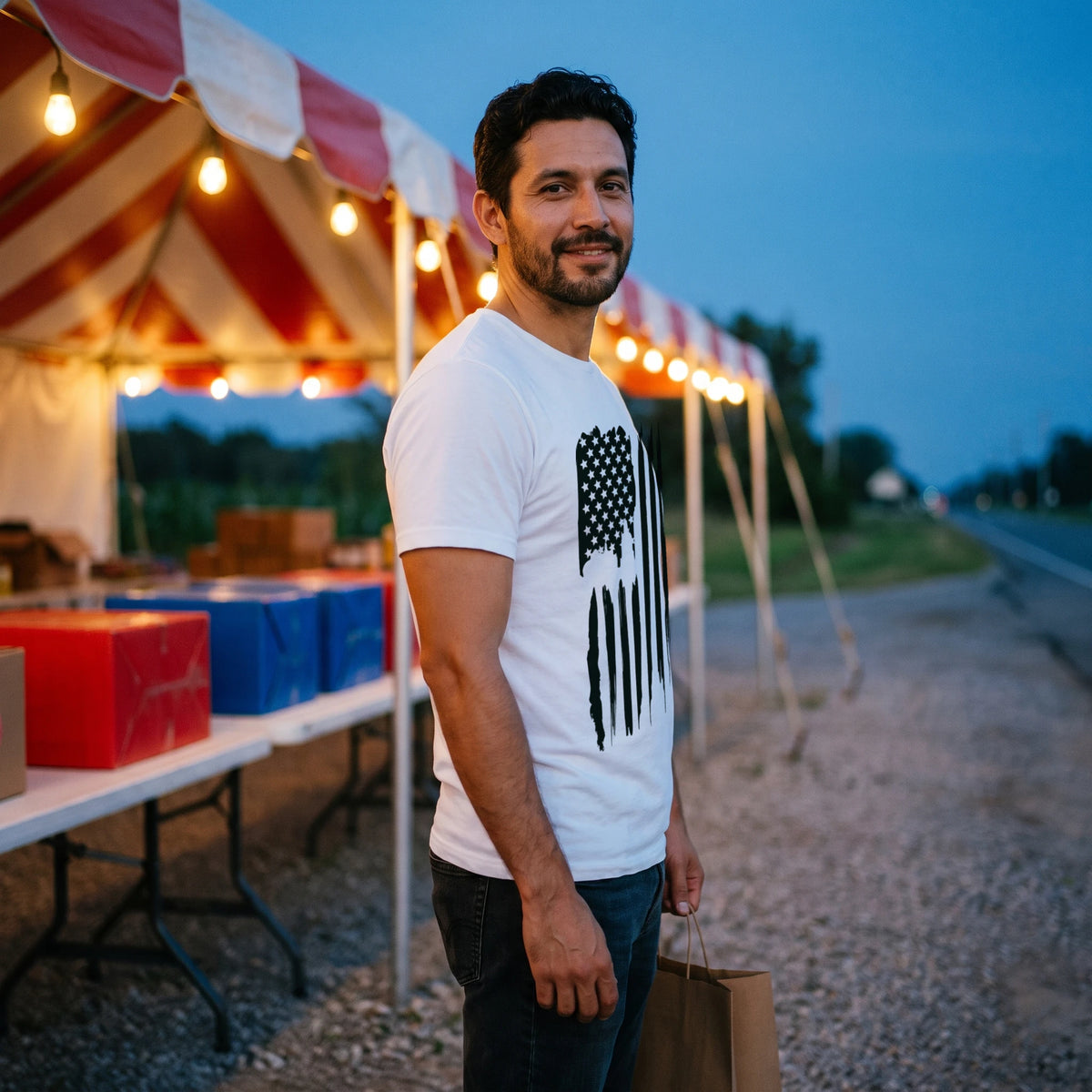 Man at a roadside fireworks stand at dusk under a striped tent and string lights, turned to the side and holding a paper bag, wearing the American Flag T-Shirt in White with the tall vertical black brush-stroke American flag printed down the front