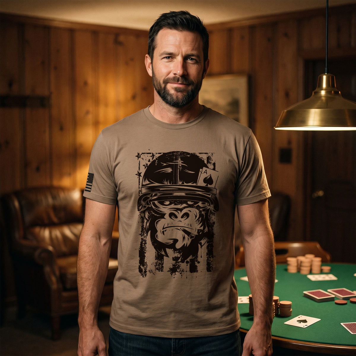 Man standing beside a green-felt card table in a wood-panelled lodge room at night, an ace of spades face up, wearing the American Gorilla Ace of Spades T-Shirt in Brown Savana with the black gorilla graphic printed large on the front