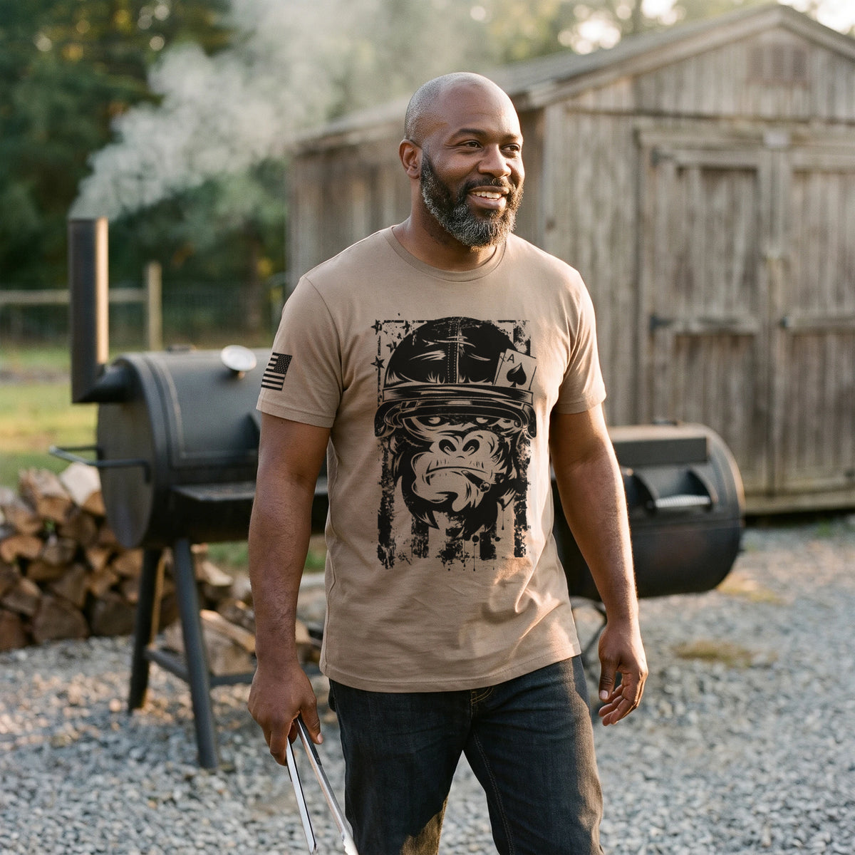 Man walking past a smoking offset BBQ smoker behind a roadside smokehouse at golden hour, tongs in hand, wearing the American Gorilla Ace of Spades T-Shirt in Brown Savana with the black gorilla graphic printed large on the front
