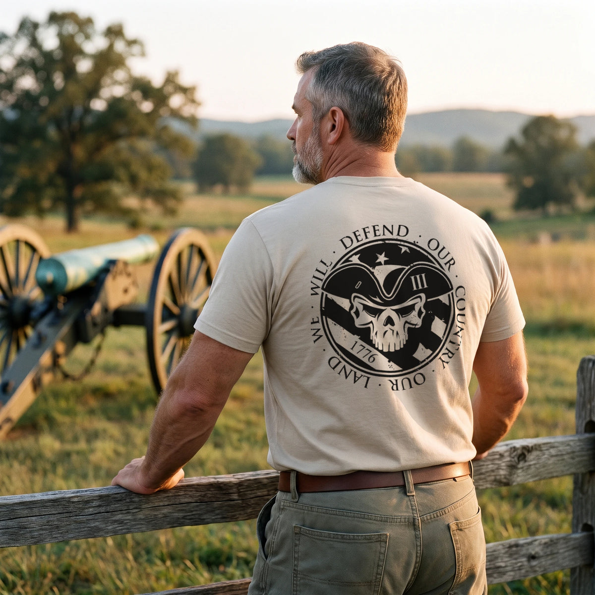 Man resting his arms on a split-rail fence at a Revolutionary War battlefield park beside an antique cannon at golden hour, wearing the American Patriot T-Shirt in Sand with the 1776 tricorn skull seal printed large on the back