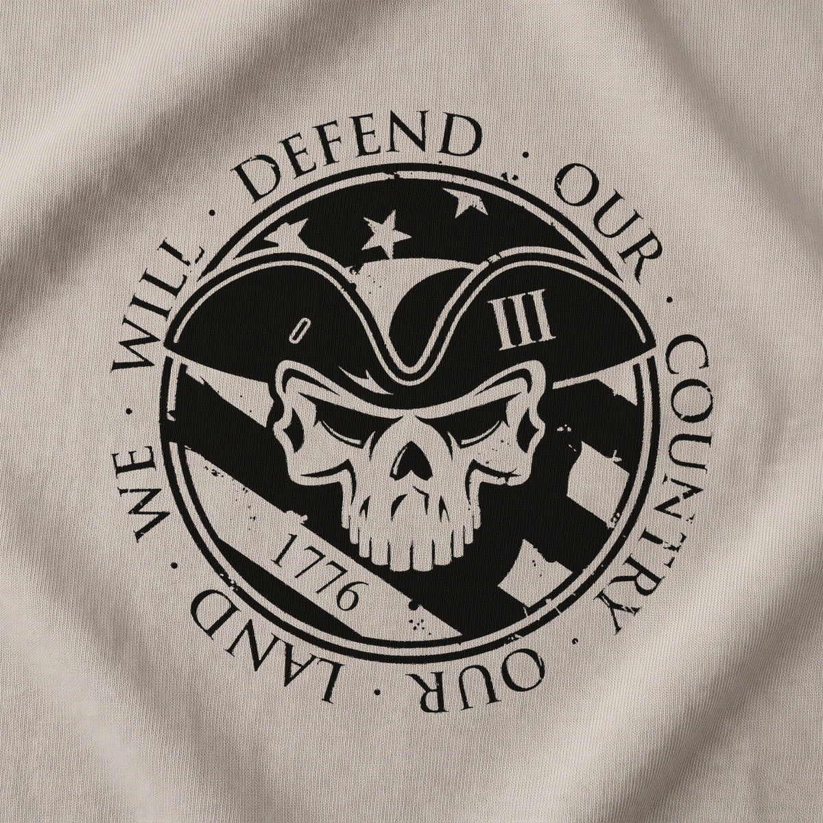 Close-up of the American Patriot T-Shirt back print on Sand: skull in a tricorn hat over the Stars and Stripes and 1776, ringed by WE WILL DEFEND OUR COUNTRY · OUR LAND (digitally composited print visualization, not a photograph)