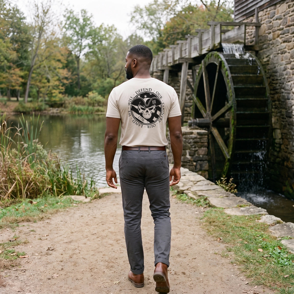 Back view of a man walking past an old stone gristmill and its wooden waterwheel, wearing the American Patriot T-Shirt in Sand with the tricorn skull seal printed on the back