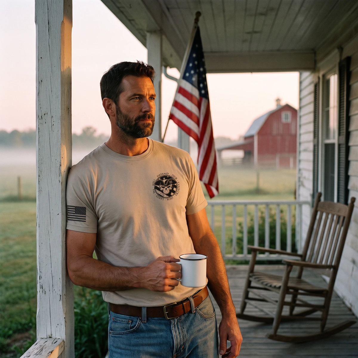 Man leaning on a farmhouse porch post at dawn with a coffee mug, an American flag behind him, wearing the American Patriot T-Shirt in Sand with the small skull seal on the left chest and an American flag on the right sleeve