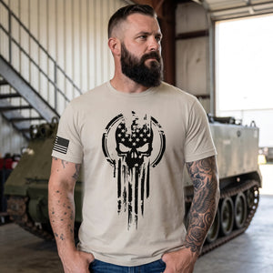 Bearded man with tattooed arms wearing the Sand American Warrior Skull T-Shirt in a hangar beside a vintage armored vehicle