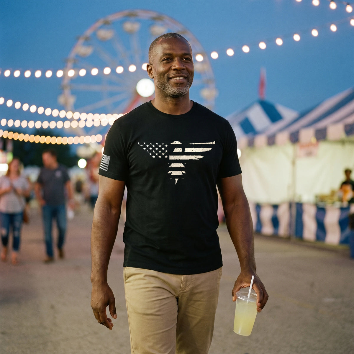 Man walking through a county fair midway at dusk with a lemonade, Ferris wheel and string lights behind him, wearing the Bald Eagle T-Shirt in Black with the stars-and-stripes eagle print across the chest and an American flag on the right sleeve