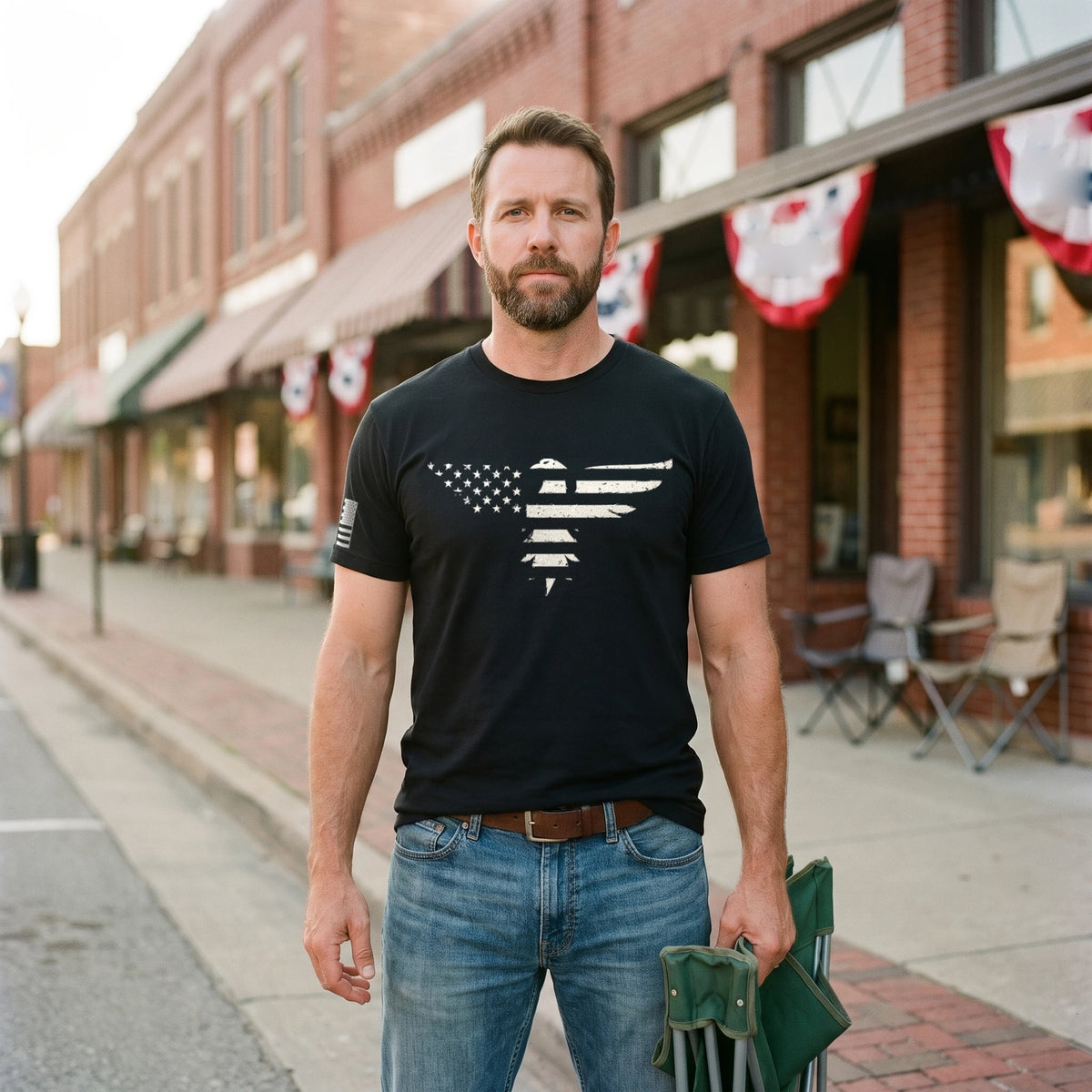 Man standing on the brick sidewalk of a small-town Main Street before a parade, red, white and blue bunting behind him, wearing the Bald Eagle T-Shirt in Black with the stars-and-stripes eagle print across the chest and an American flag on the right sleeve