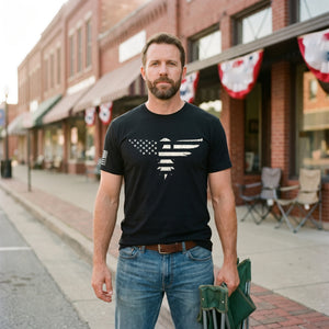 Man standing on the brick sidewalk of a small-town Main Street before a parade, red, white and blue bunting behind him, wearing the Bald Eagle T-Shirt in Black with the stars-and-stripes eagle print across the chest and an American flag on the right sleeve