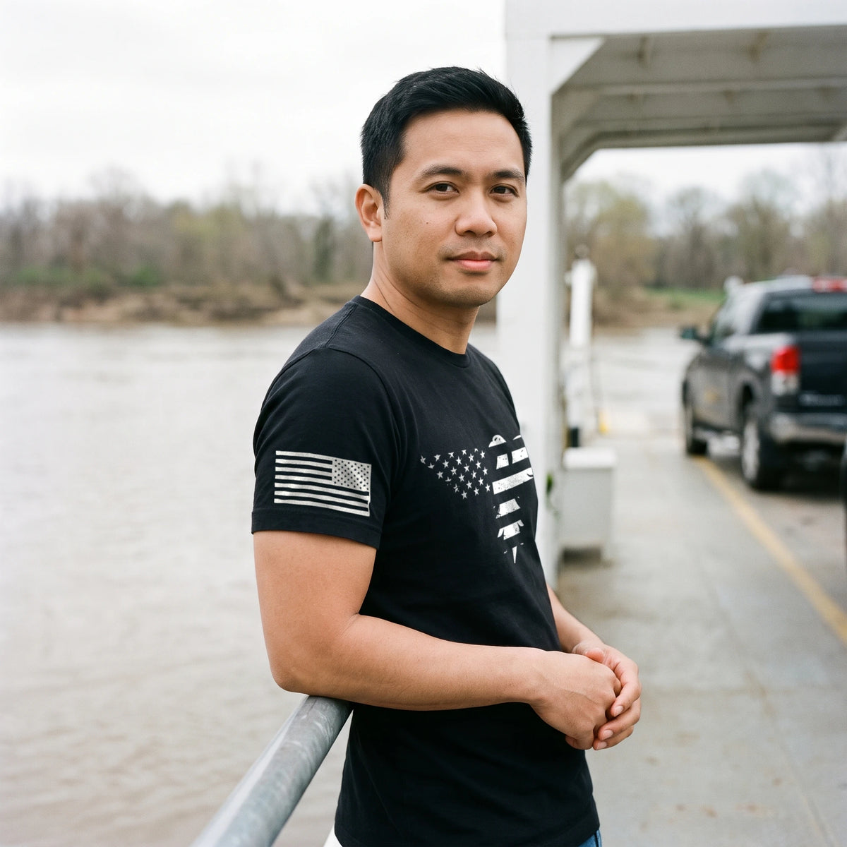 Man leaning on the rail of a small river car ferry on an overcast morning, wide river behind him, wearing the Bald Eagle T-Shirt in Black with the stars-and-stripes eagle print across the chest and an American flag on the right sleeve
