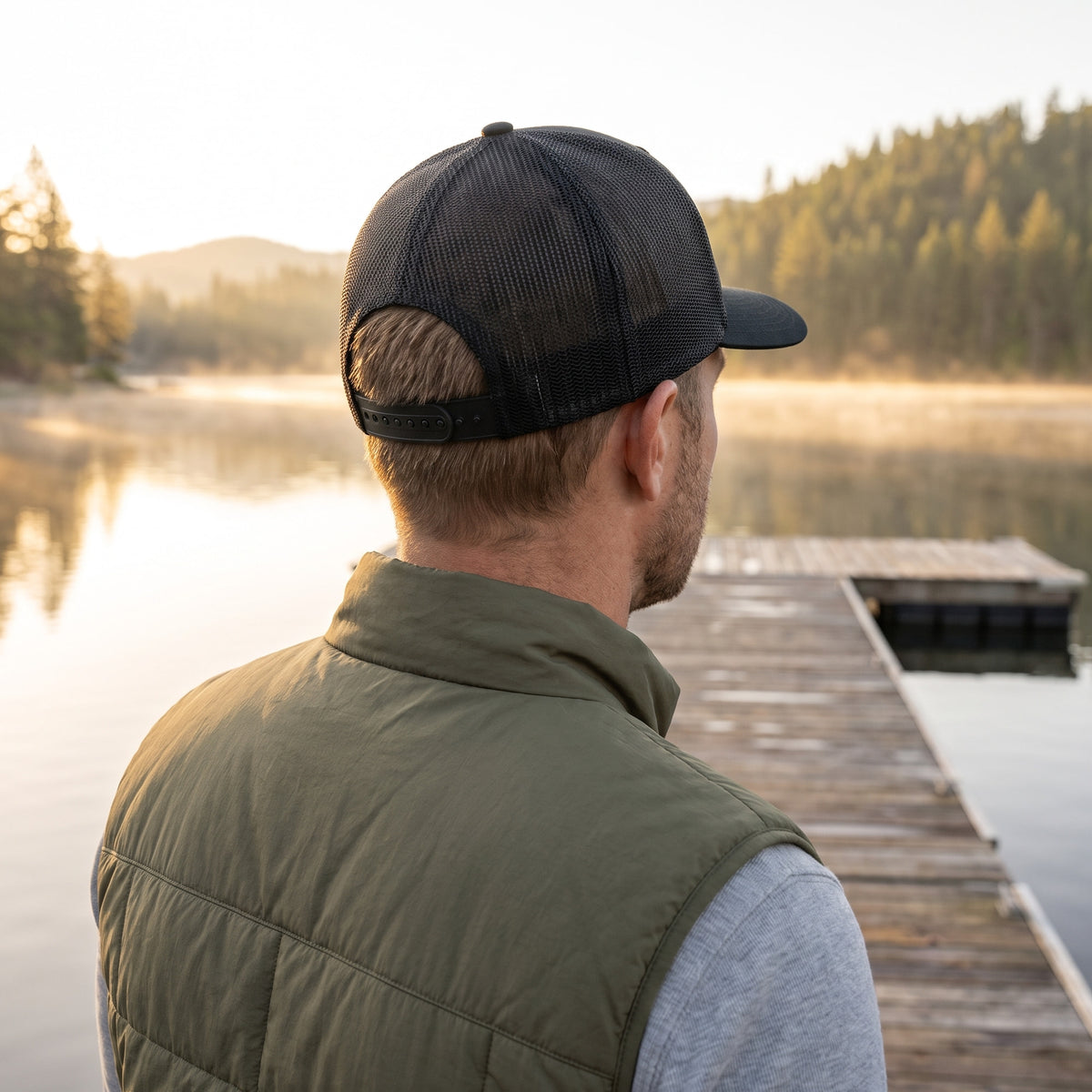 Back view of the Crusader Cross trucker hat on a lake dock at sunrise showing the black mesh back and plastic snapback