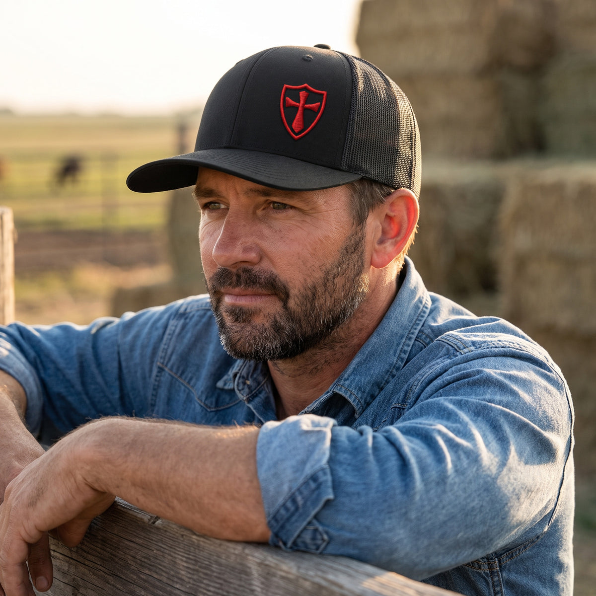 Rancher wearing the black and red Crusader Cross trucker hat beside hay bales at golden hour