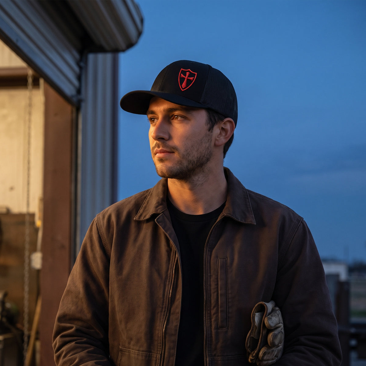 Man outside a fabrication workshop at dusk wearing the black and red Crusader Cross trucker hat