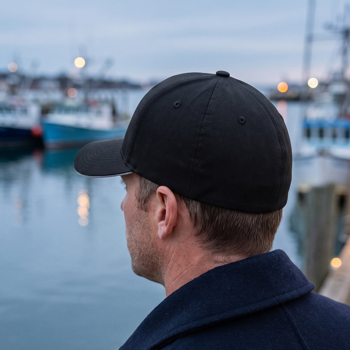 Rear three-quarter view of the black Crusader Cross stretch fit cap on a man on a harbor pier: clean, closed stretch-fit back with no embroidery