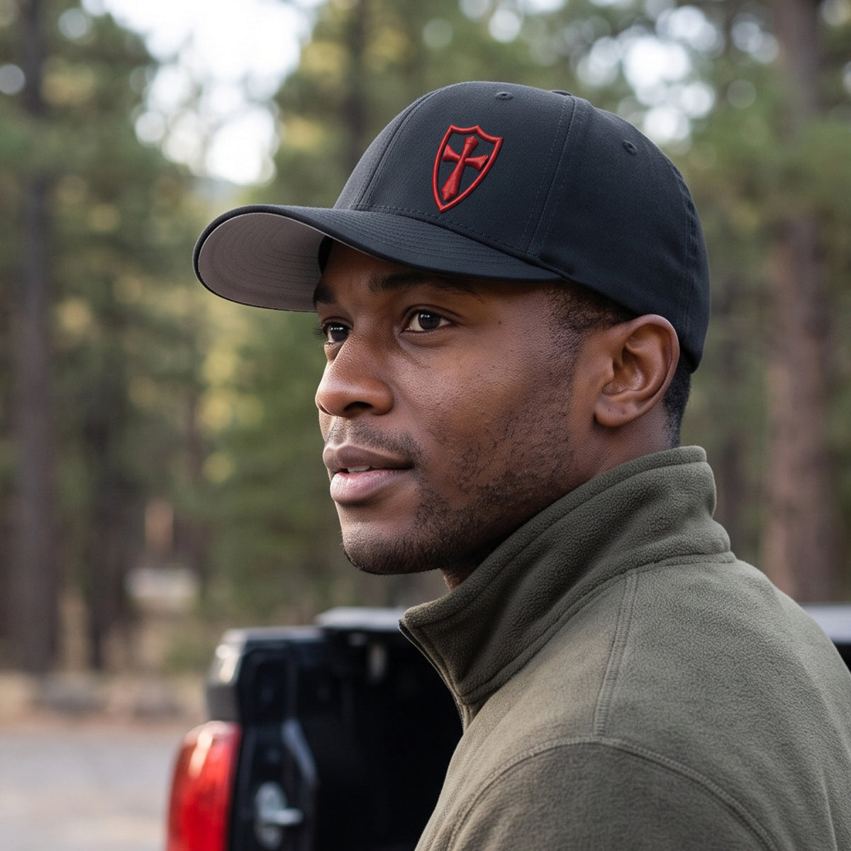 Man beside a pickup at a forest trailhead wearing the black Crusader Cross stretch fit cap: side three-quarter view of the crown, curved bill and red 3D puff Crusader shield