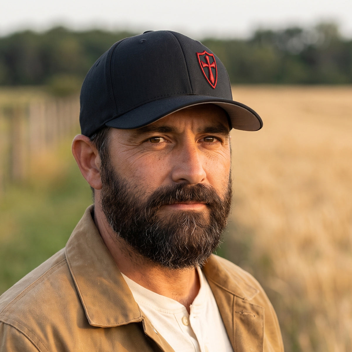 Bearded man at the edge of a wheat field wearing the black Crusader Cross stretch fit cap with the red 3D puff Crusader shield, front three-quarter view