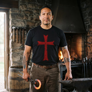Man with long dark hair standing at a stone blacksmith forge by the fire, holding tongs with a glowing horseshoe, wearing the Crusader Cross T-Shirt in Black with the red distressed Crusader cross on the front