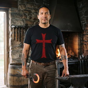 Man with long dark hair standing at a stone blacksmith forge by the fire, holding tongs with a glowing horseshoe, wearing the Crusader Cross T-Shirt in Black with the red distressed Crusader cross on the front