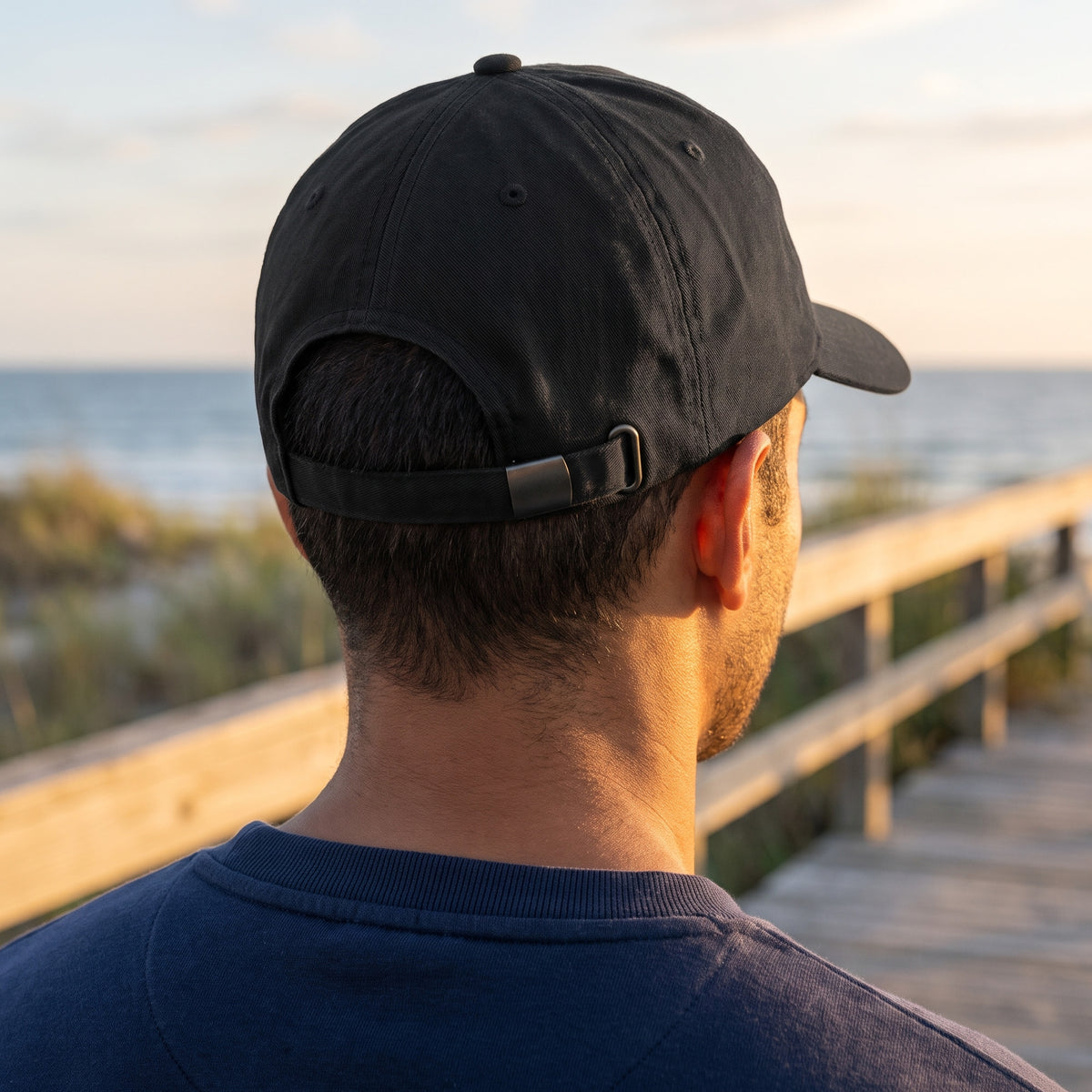Back view of the black Dion Wear unstructured cap on a coastal boardwalk at sunset, showing the plain back and cloth hideaway strap