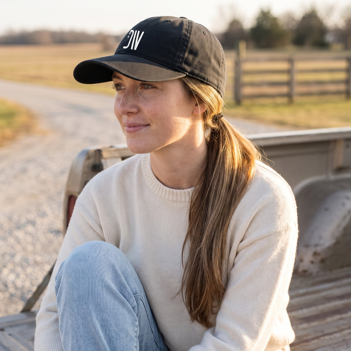 Woman sitting on a pickup tailgate on a country road at golden hour wearing the black Dion Wear unstructured cap