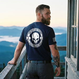 Man on the catwalk of a mountain fire lookout tower at dawn, looking back over his shoulder, wearing the Divided We Fall Skull T-Shirt in Navy: United We Stand, Divided We Fall skull seal printed on the back, American flag on the right sleeve