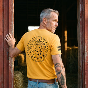 Man with grey stubble standing in a red barn doorway at sunrise, looking back over his shoulder, wearing the Don't Tread on Me T-Shirt in Gold: Gadsden rattlesnake seal printed on the back, American flag on the right sleeve