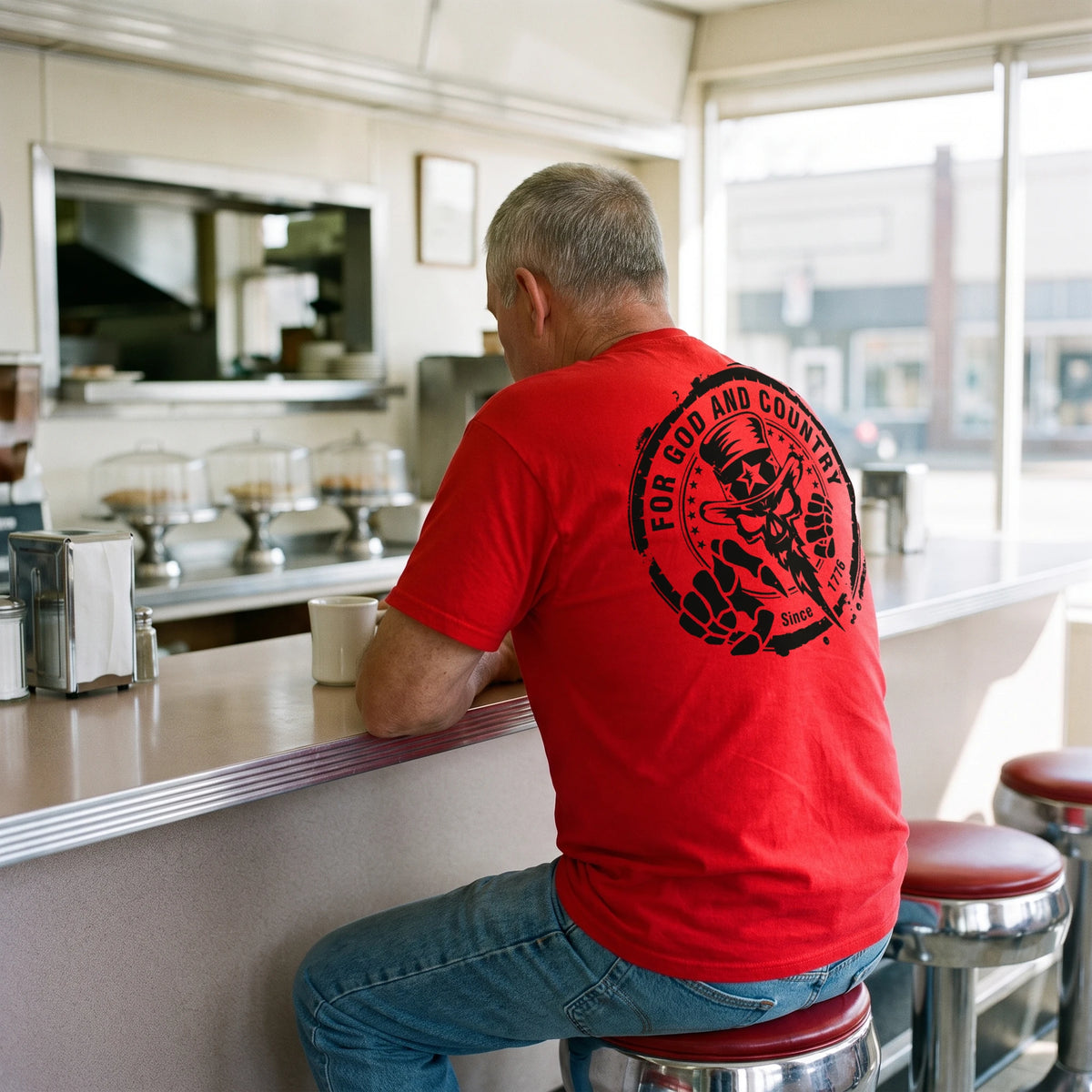 Back view of the For God and Country T-Shirt in Red on an older man seated at a small-town diner counter: For God and Country seal (Uncle Sam skull, Since 1776) printed large on the upper back