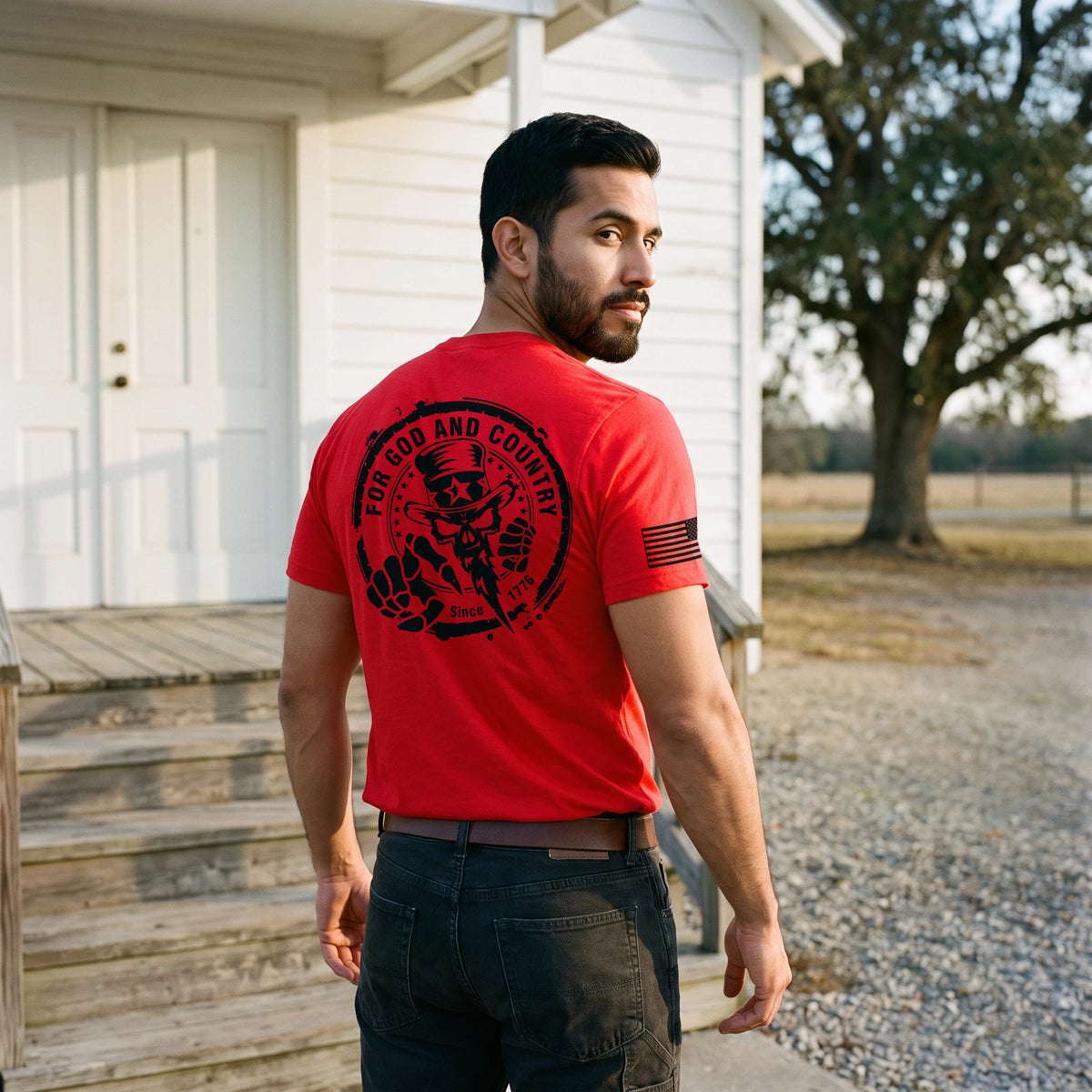Man looking back over his shoulder at the steps of a white country chapel in early sunlight, wearing the For God and Country T-Shirt in Red: For God and Country seal (Uncle Sam skull, Since 1776) printed large on the back, black American flag on the right sleeve