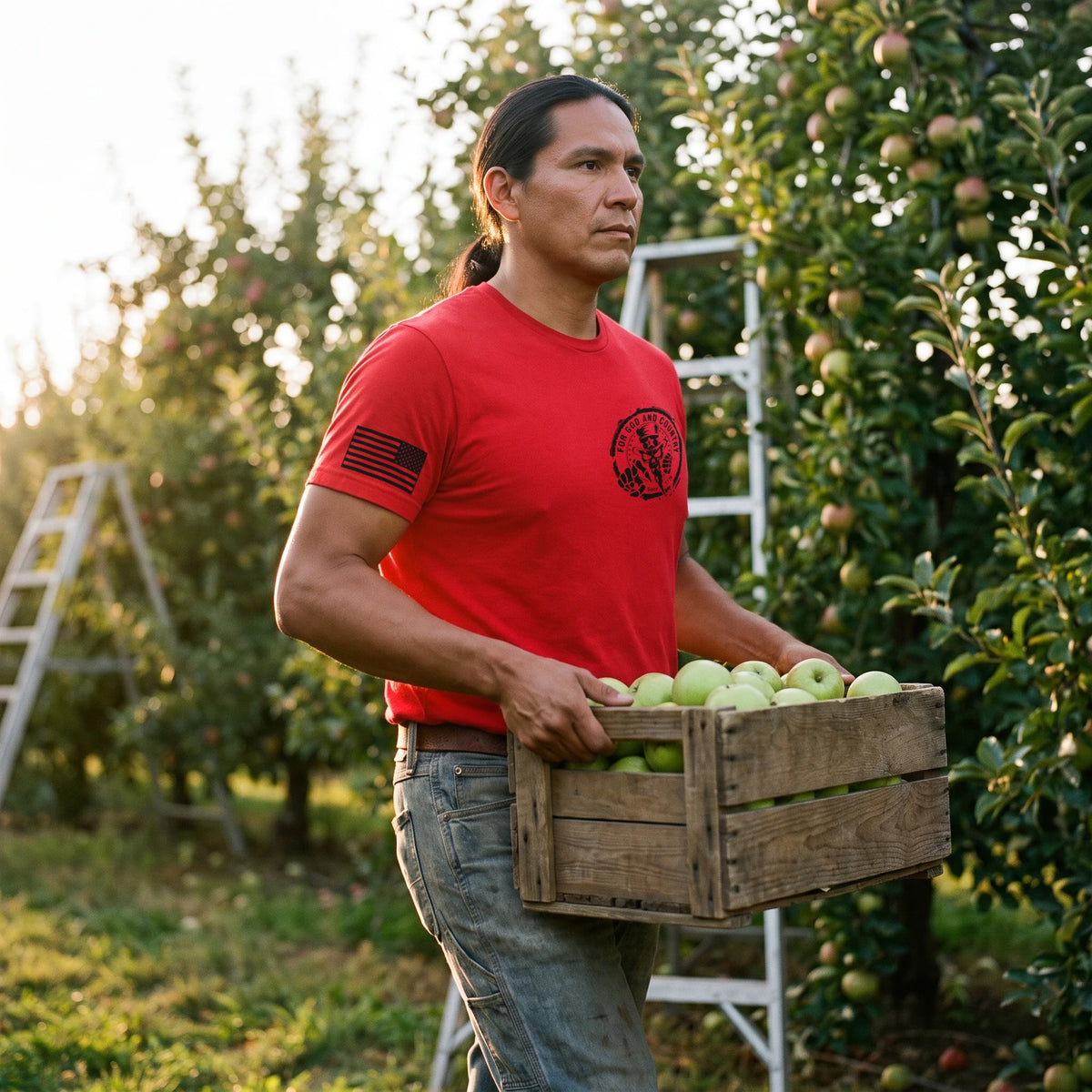 Man carrying a crate of green apples through an orchard, wearing the For God and Country T-Shirt in Red with the small For God and Country seal (Uncle Sam skull, Since 1776) on the left chest and a black American flag on the right sleeve