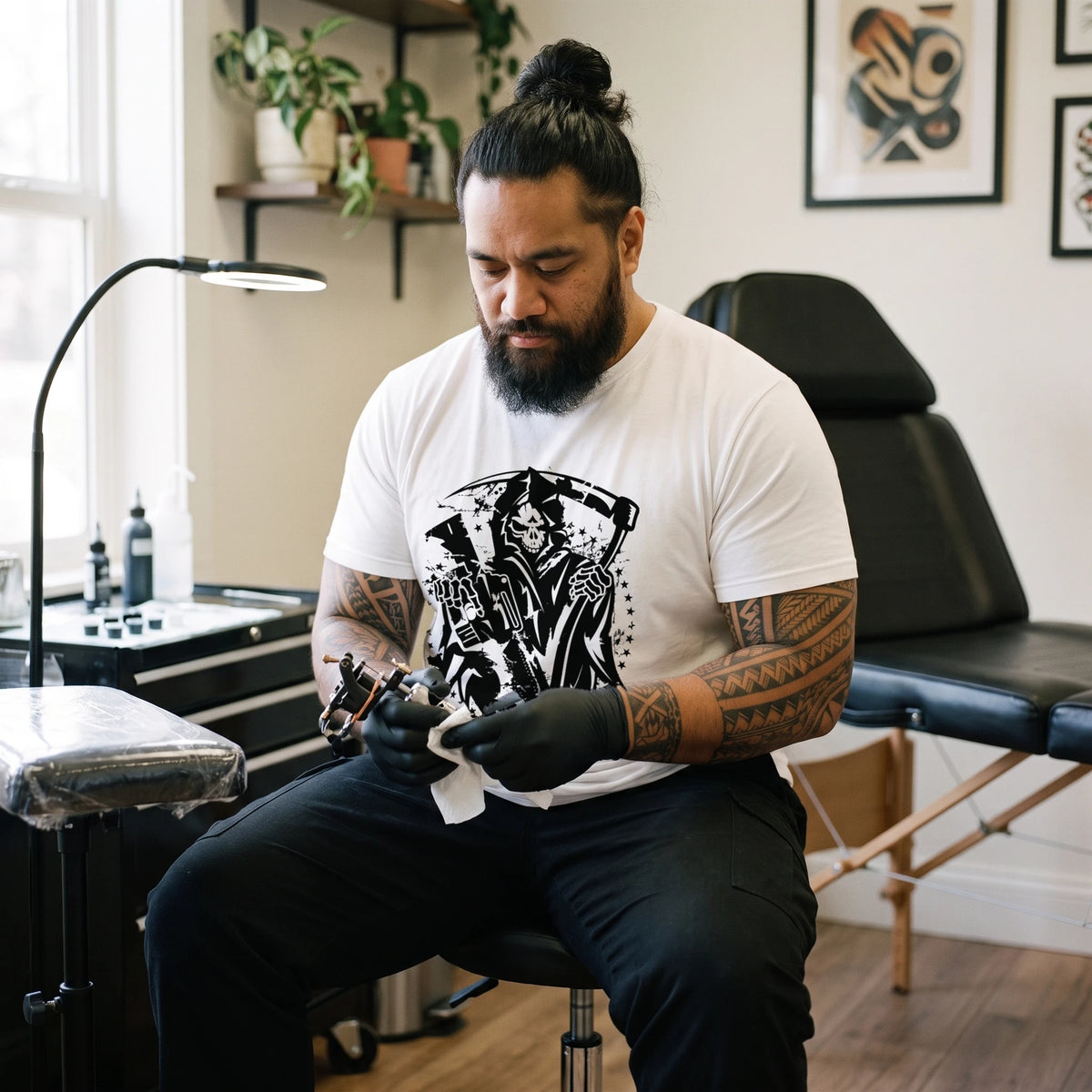 Tattoo artist with tattooed forearms cleaning his machine at his studio workstation, wearing the Grim Reaper T-Shirt in White with the black Grim Reaper with a scythe, a rifle, stars and splatter printed large across the front