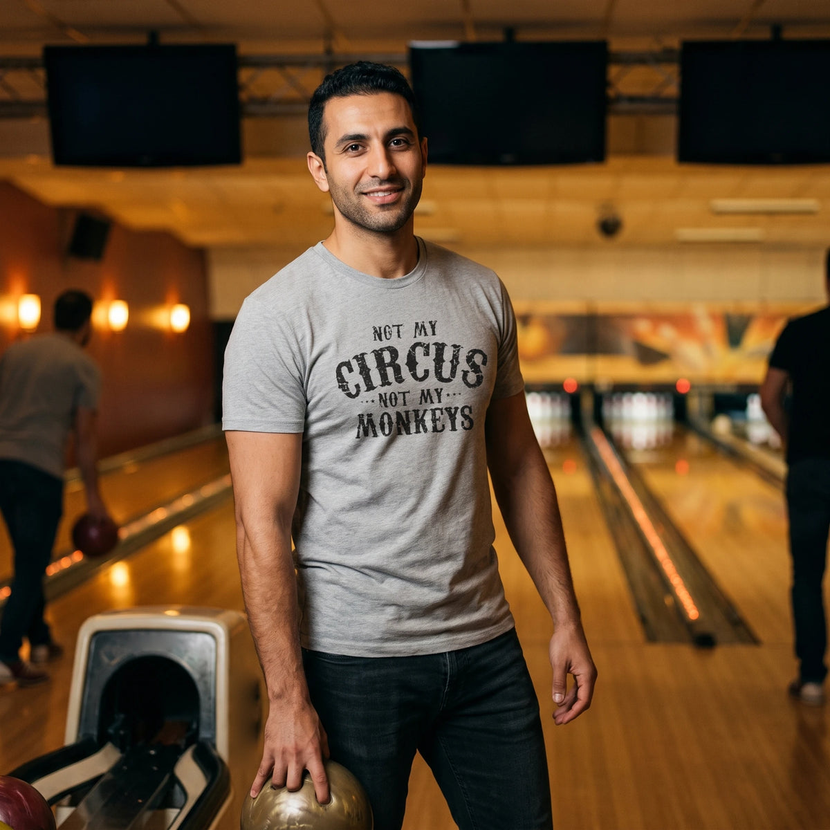 Smiling man holding a bowling ball by the ball return of a retro bowling alley, wearing the Not My Circus, Not My Monkeys T-Shirt in Heather Gray with black distressed circus-poster lettering reading Not My Circus, Not My Monkeys printed large across the chest
