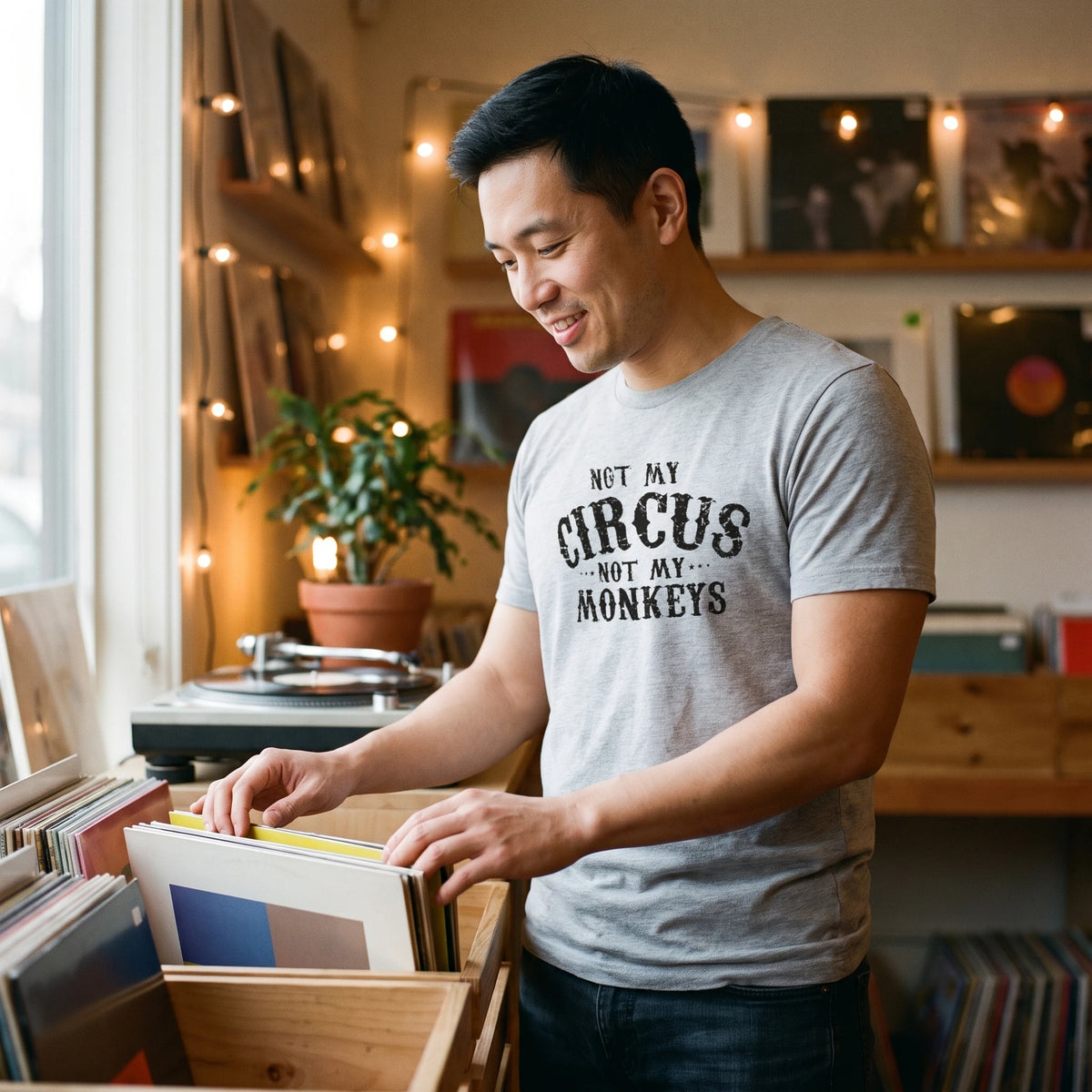 Man flipping through vinyl records in an independent record shop, wearing the Not My Circus, Not My Monkeys T-Shirt in Heather Gray with black distressed circus-poster lettering reading Not My Circus, Not My Monkeys printed large across the chest