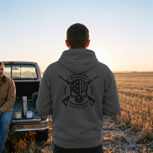 Man in the charcoal One Nation Under God Hoodie at a pickup tailgate in a harvested wheat field at dawn, seen from behind with the black crest across the back