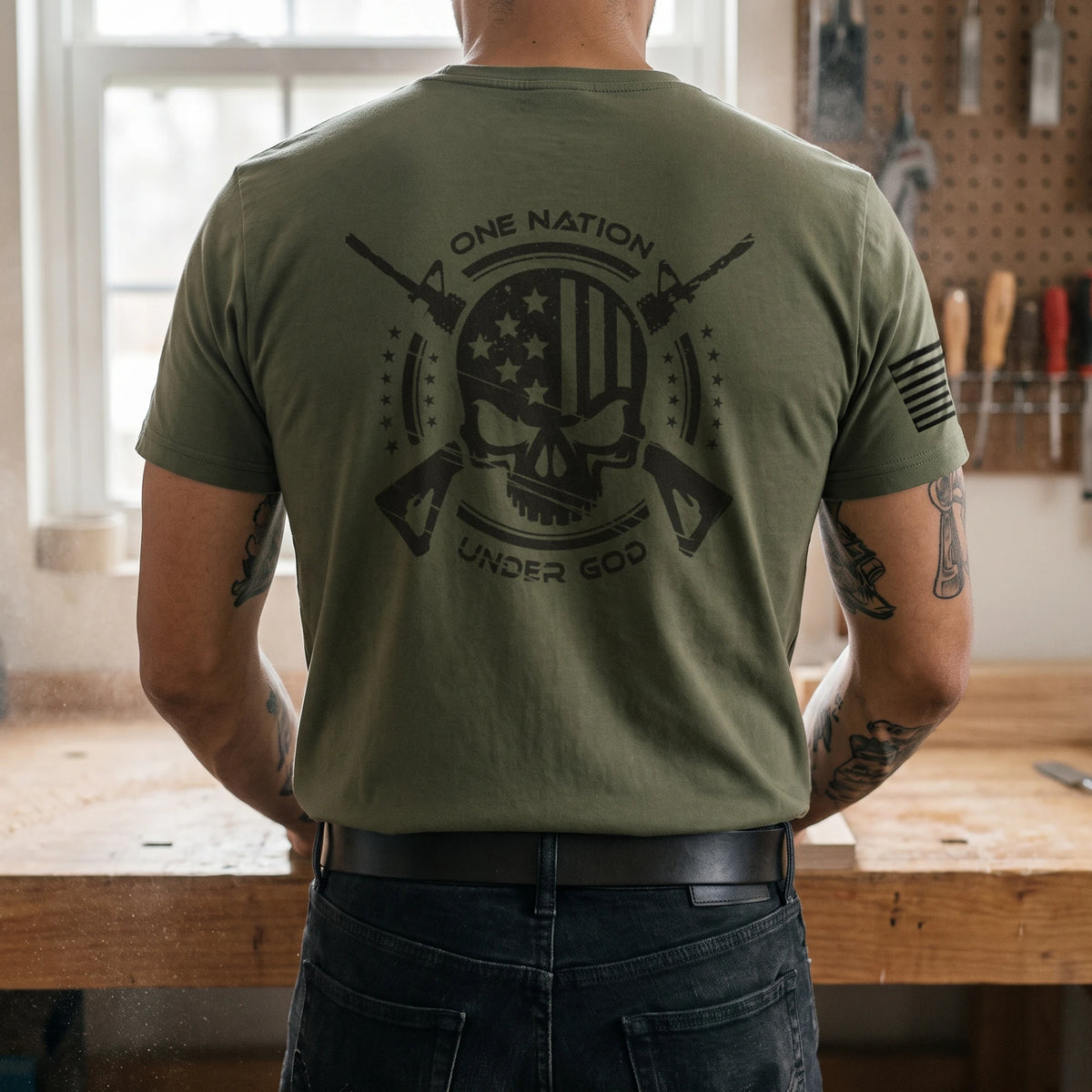 Back view of the One Nation Under God T-Shirt in Military Green worn in a woodshop: skull and American flag back print with crossed rifles and stars