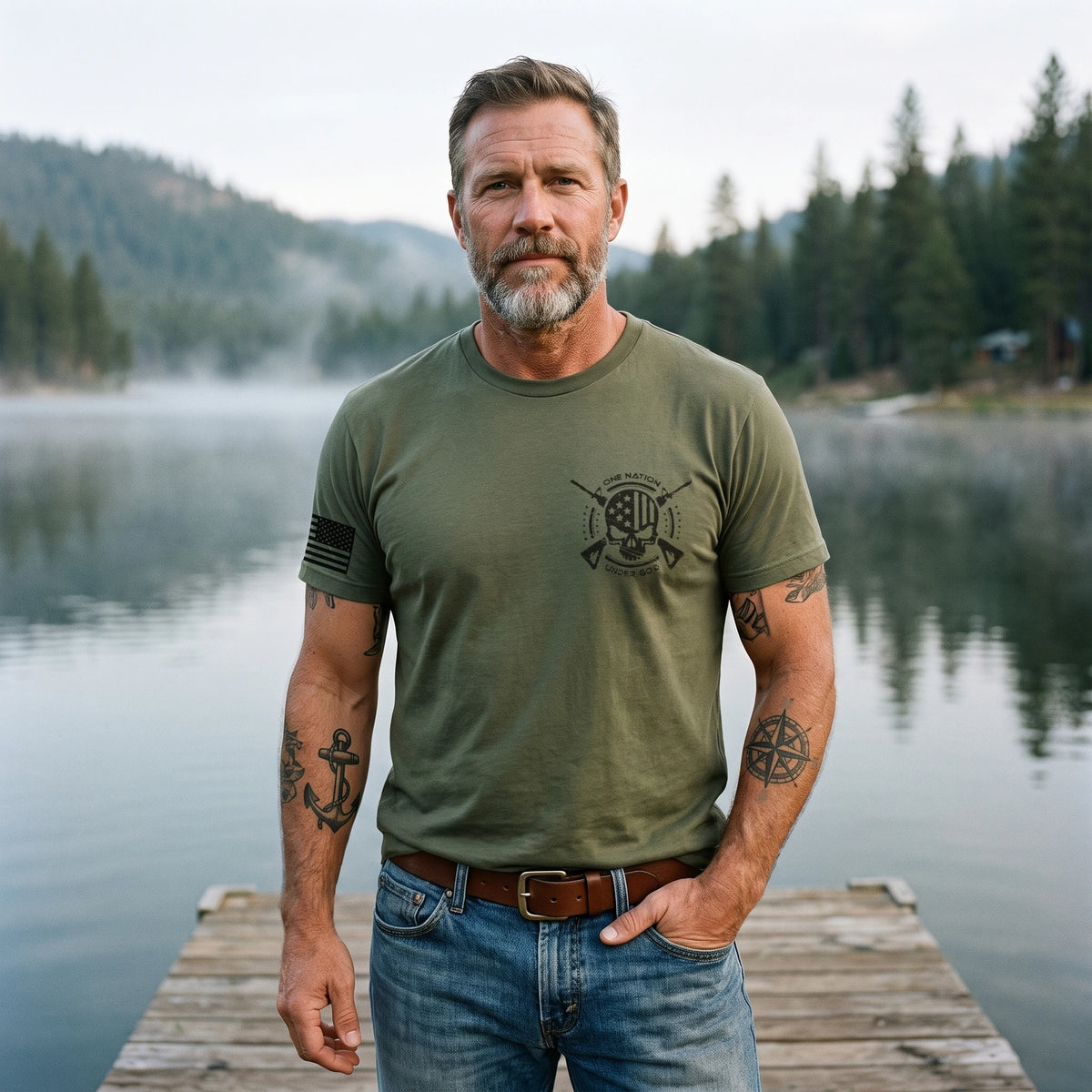 Man with a salt-and-pepper beard and tattooed forearms on a lakeside dock wearing the One Nation Under God T-Shirt in Military Green: small left-chest emblem and American flag on the right sleeve