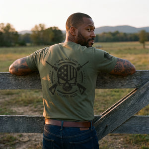 Bearded man with tattooed arms leaning on a wooden ranch gate at dusk, looking over his shoulder, wearing the One Nation Under God T-Shirt in Military Green with the large back print
