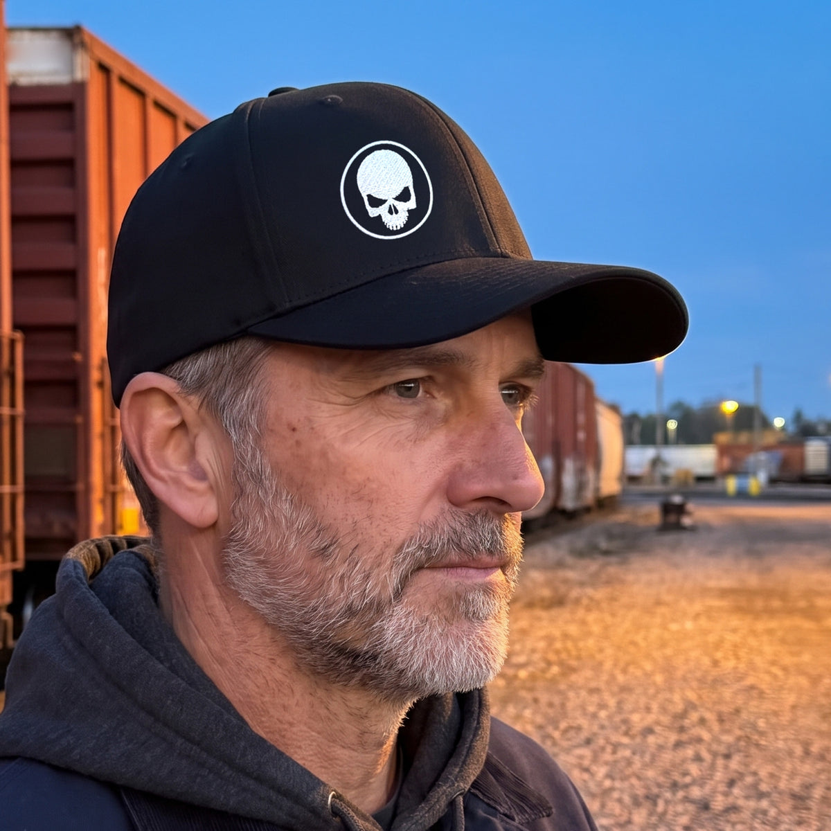 Man in a freight rail yard at dusk wearing the black Skull Stretch Fit Cap: side three-quarter view of the crown, curved bill and embroidered skull