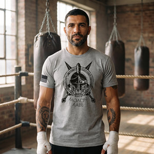 Man with a dark beard and tattooed forearms in an old boxing gym wearing the Spartan Molon Labe T-Shirt in Heather Gray: Spartan helmet and MOLON LABE front print, American flag on the right sleeve