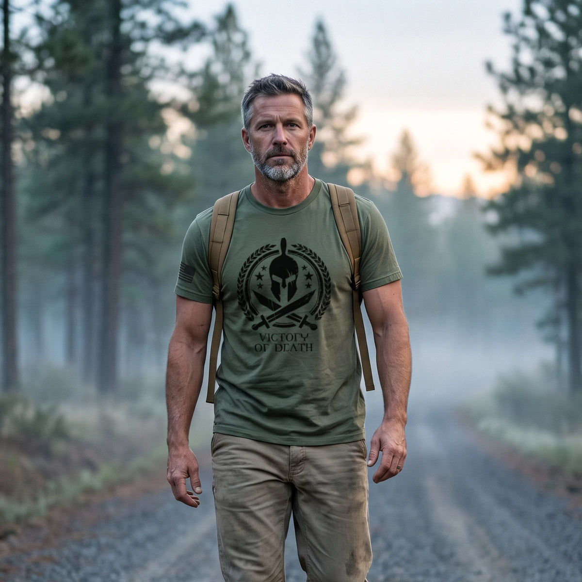 Man rucking along a misty pine forest road at dawn with a plain tan rucksack, wearing the Spartan Warrior T-Shirt in Military Green with the Victory or Death print on the front and an American flag on the right sleeve