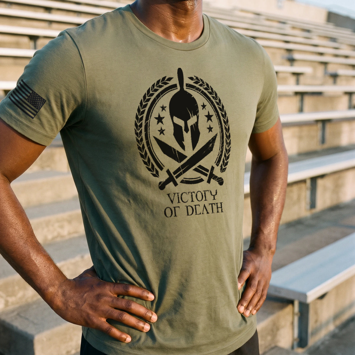 Close view of a man with his hands on his hips on concrete stadium stairs at sunrise, wearing the Spartan Warrior T-Shirt in Military Green with the Spartan helmet, laurel wreath and Victory or Death print on the chest and an American flag on the right sleeve