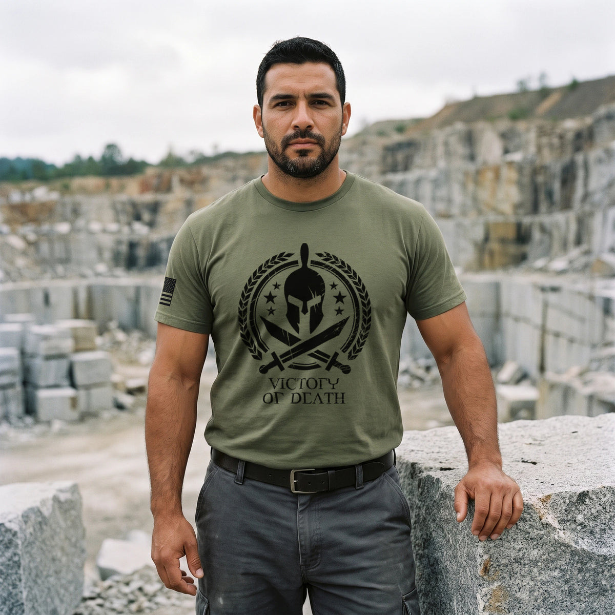 Man resting a hand on a cut granite block at the edge of a stone quarry, wearing the Spartan Warrior T-Shirt in Military Green with the Victory or Death Spartan helmet print on the front and an American flag on the right sleeve