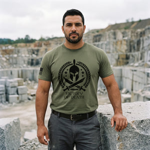 Man resting a hand on a cut granite block at the edge of a stone quarry, wearing the Spartan Warrior T-Shirt in Military Green with the Victory or Death Spartan helmet print on the front and an American flag on the right sleeve