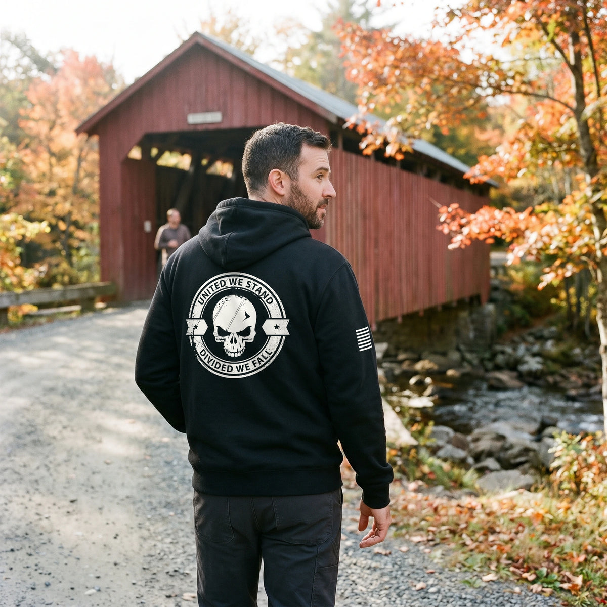Man in the black United We Stand Hoodie walking from a red covered bridge in autumn, with the back print and the American flag on the right sleeve